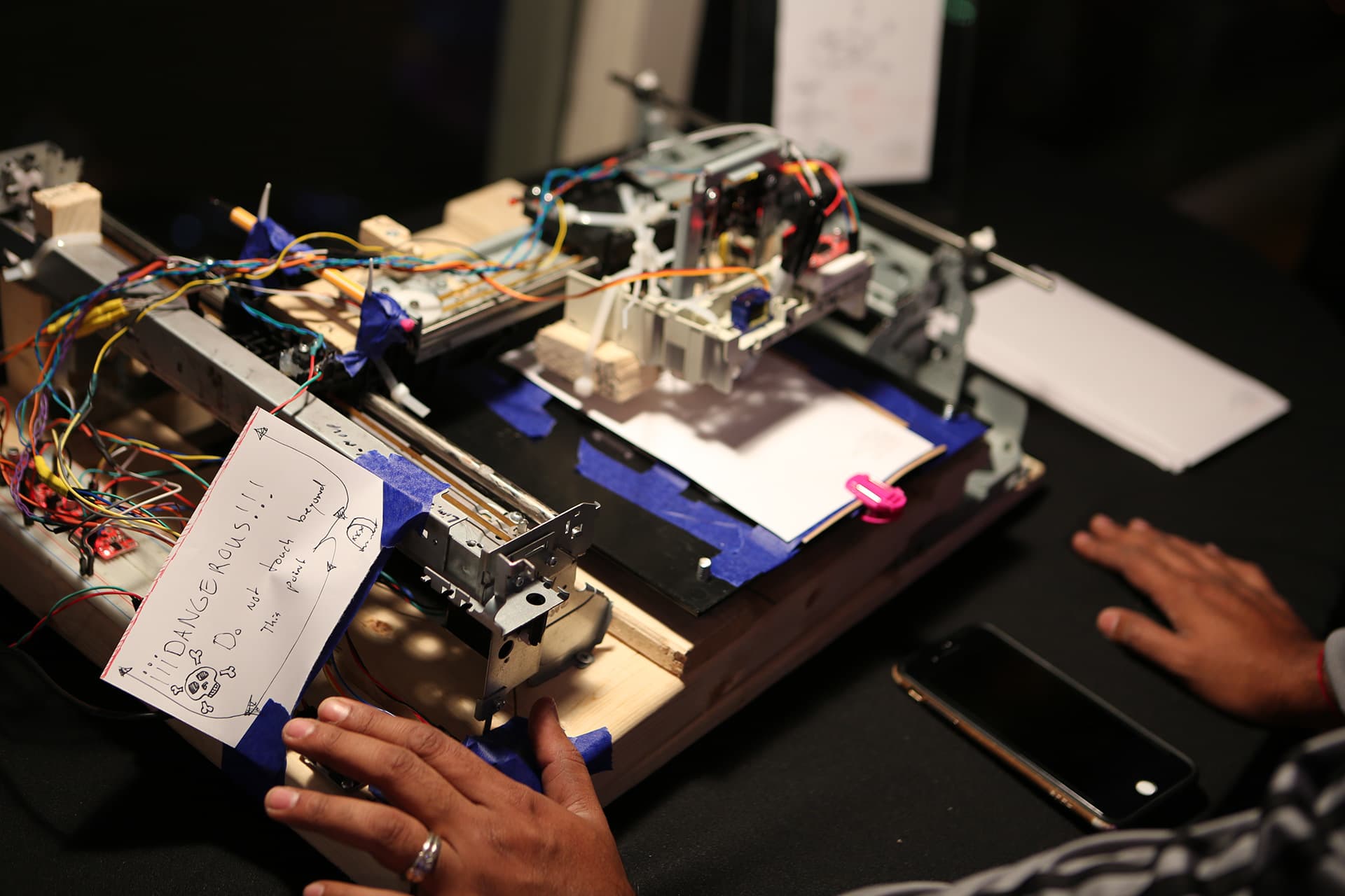 Custom-built perler-bead plotting machine on a desk: breadboard, colored wiring, blue painter's tape, and a hand guiding the rig